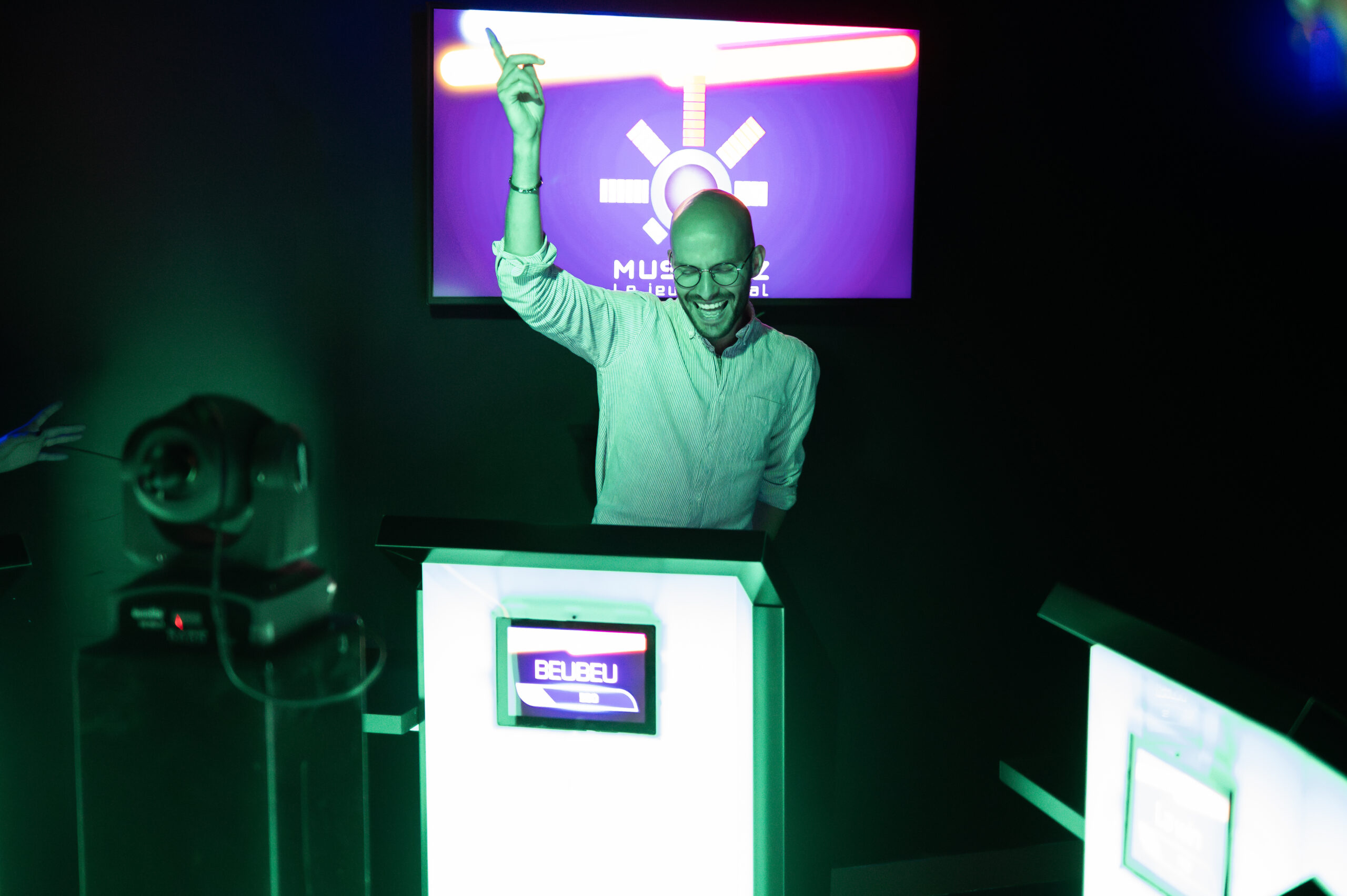 Homme qui joue à une session Musi'Quiz, il est derrière son pupitre. Il est joyeux car il a obtenu une bonne réponse, on le voit grâce au pupitre ainsi que la salle qui s'éclair en vert.