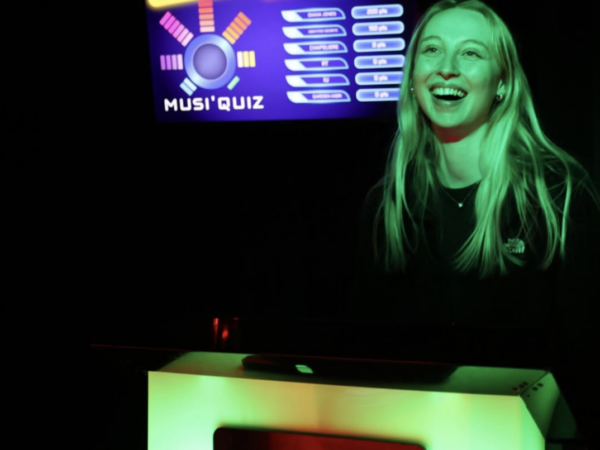 Photographie d'une fille qui joue dans une salle Musi'Quiz, elle est derrière un pupitre Musi'Quiz. Elle rigole car elle a obtenu une bonne réponse. On le remarque à la lumière verte qui éclaire la pièce.
