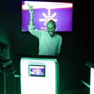 Photographie d’un homme qui joue à Musi’Quiz. Il se tient debout derrière son pupitre. Il exulte de joie, le doigt pointé vers le ciel, après avoir obtenu une bonne réponse. Son pupitre est allumé en vert.